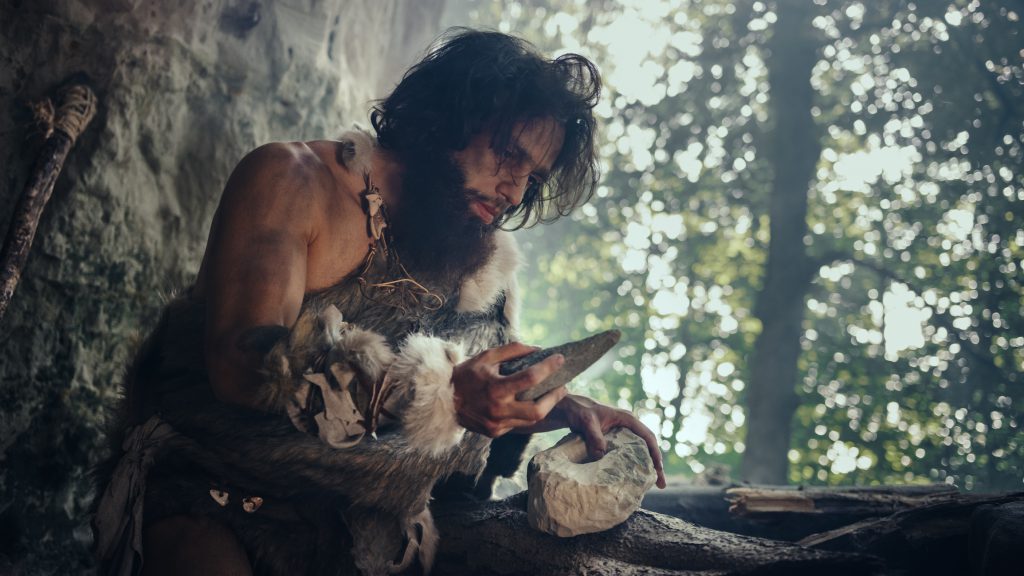 Primeval Caveman Wearing Animal Skin Holds Sharp Stone and Makes First Primitive Tool for Hunting Animal Prey, or to Handle Hides. Neanderthal Using Handax. Dawn of Human Civilization ERP(Enterprise Resource Planning) System in Singapore